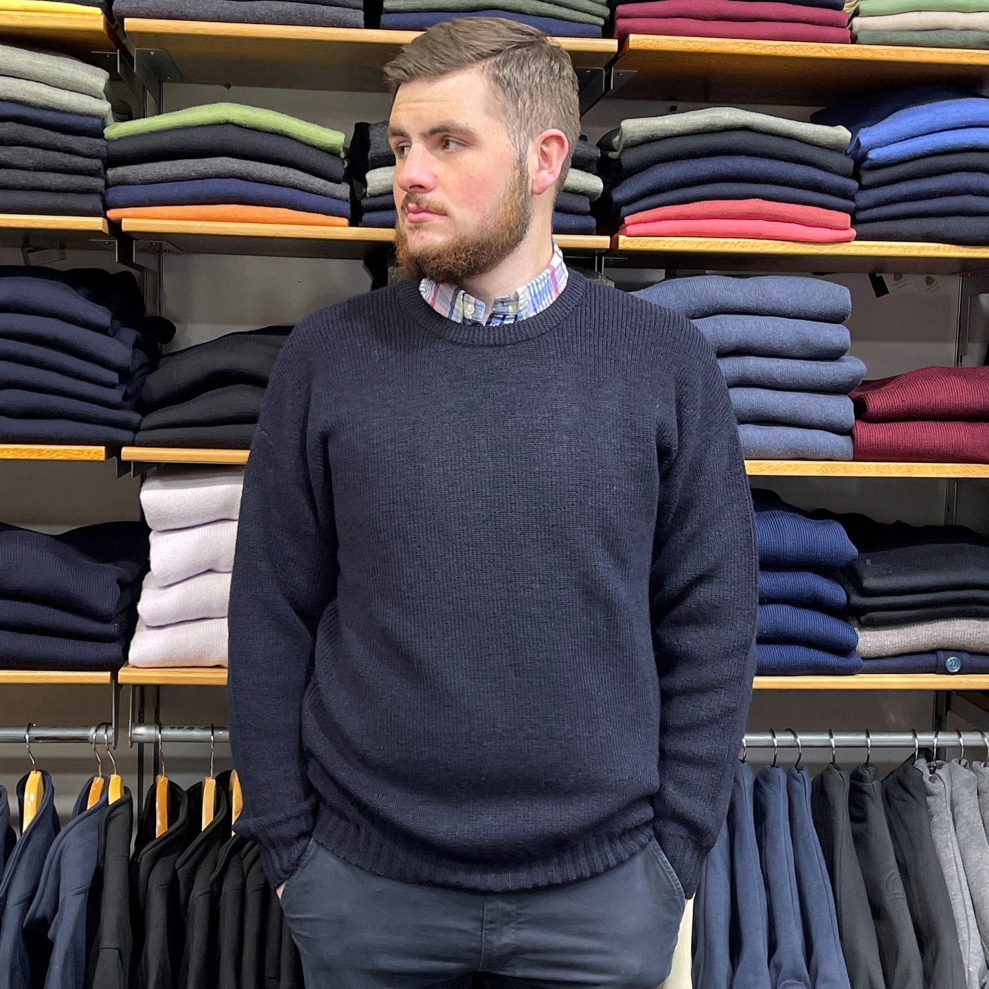 A man wearing a navy sweater, showcasing The Ultimate Men's Winter Wardrobe for 2025, with folded clothes in the background.