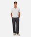 A man modeling a white short-sleeve polo shirt and dark gray drawstring trousers with beige slip-on shoes, standing against a light gray studio background.