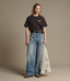 A model stands against a plain beige studio background wearing a black short-sleeve T-shirt with a small white chest logo, wide-leg blue jeans, a black belt, and holding a light plaid shirt under one arm.