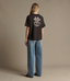 A woman stands with her back to the camera wearing a black t-shirt with a large white graphic print on the back and wide-leg blue jeans, photographed in a studio setting with a solid beige background.