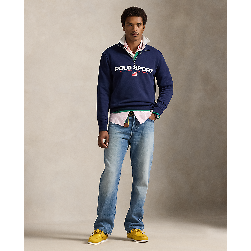 A male model stands against a textured beige studio backdrop wearing a navy blue Polo Sport quarter-zip sweatshirt over a white collared shirt, blue jeans, and yellow shoes. The Polo Sport logo is clearly visible on the chest.