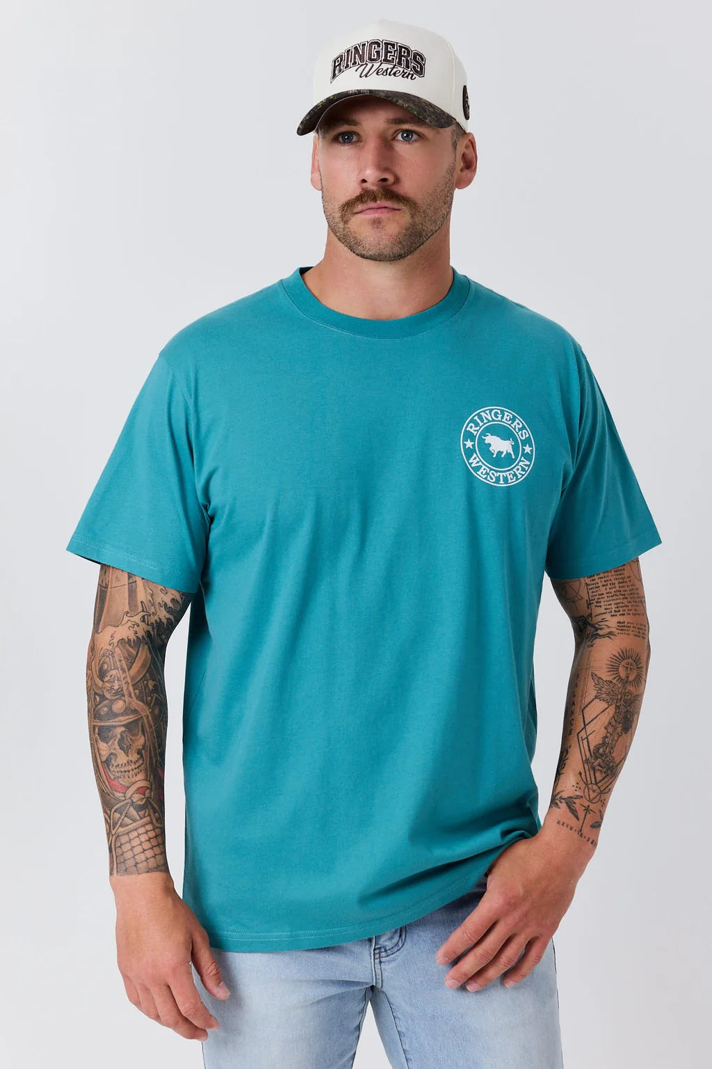A man wearing a teal short-sleeve t-shirt with a circular white graphic on the left chest, paired with a white cap and light blue jeans, standing against a light gray background.