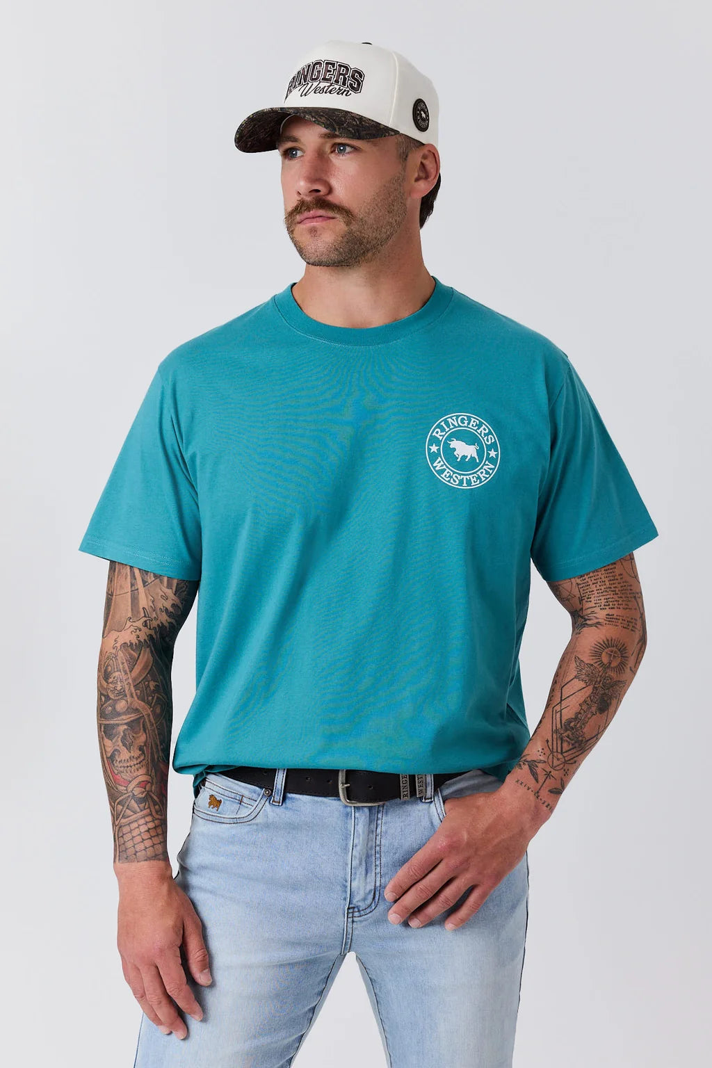 A man wearing a teal short-sleeve t-shirt with a circular white chest graphic, paired with light blue jeans and a white baseball cap, standing against a light gray studio background.