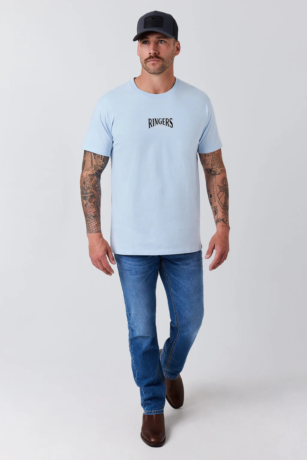 A man modeling a light blue crewneck t-shirt with the brand name 'RINGERS' printed in black on the chest, paired with blue jeans and a black baseball cap, against a plain light gray background.