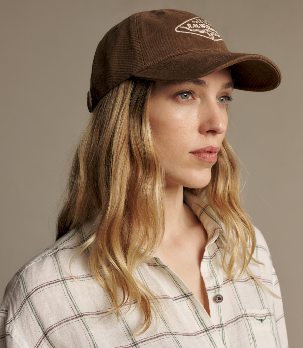 A woman wearing a brown baseball cap with a white embroidered logo and a cream plaid button-up shirt, photographed against a plain taupe background.