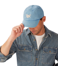 R.M.Williams - RMW Cursive Twill Cap - Blue Denim worn by man adjusting cap, showcasing embroidered logo and denim shirt.