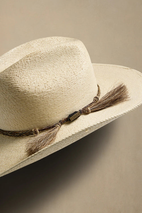 A close-up of a woven straw cowboy hat in a light beige color, featuring a brown braided band with decorative beads and two brown tassels, set against a solid neutral beige background.