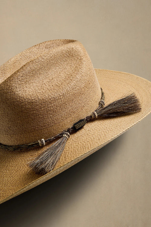 A close-up of a woven straw fedora hat in a light tan color, featuring a dark brown braided band with silver-toned beads and a matching brown tassel, set against a solid beige background.