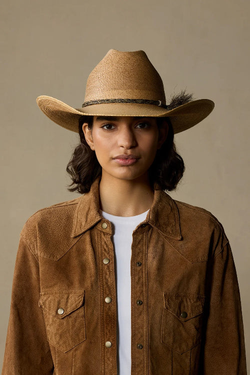 A model wearing a tan cowboy hat and a brown suede button-up shirt jacket over a white t-shirt, photographed against a solid warm beige background.