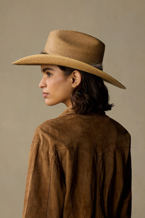 A woman shown from the back and side profile wearing a tan cowboy hat and a brown suede jacket against a plain beige studio background.