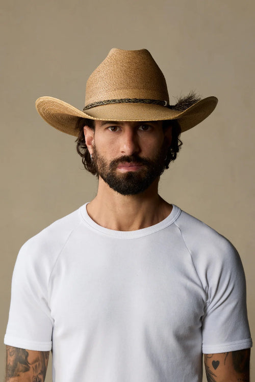 A man with a beard and tattoos wearing a light brown straw cowboy hat and a white raglan t-shirt, photographed against a solid beige background.
