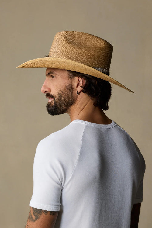 A man in profile wearing a light brown woven cowboy hat and a white short-sleeve raglan t-shirt, photographed against a solid warm beige studio background.