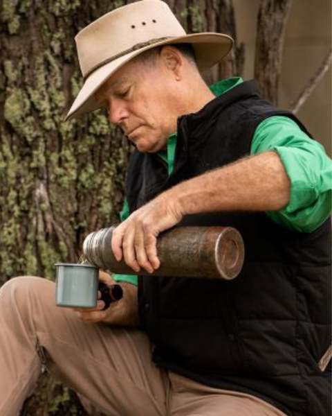 A man in a hat pouring a beverage from a thermos into a blue mug while seated outdoors.