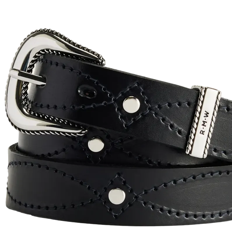A black leather belt with decorative whipstitching, silver-tone metal studs, and a silver-tone buckle, coiled on a light beige background. The end of the belt is engraved with 'R.