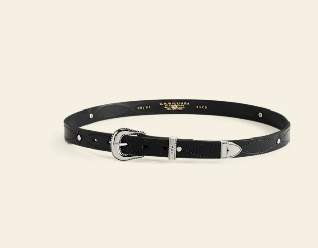 A black leather belt with silver-tone hardware, including a decorative buckle, a silver-toned keeper, and silver studs, displayed on a light cream background. The inside of the belt features gold branding text for 'B.
