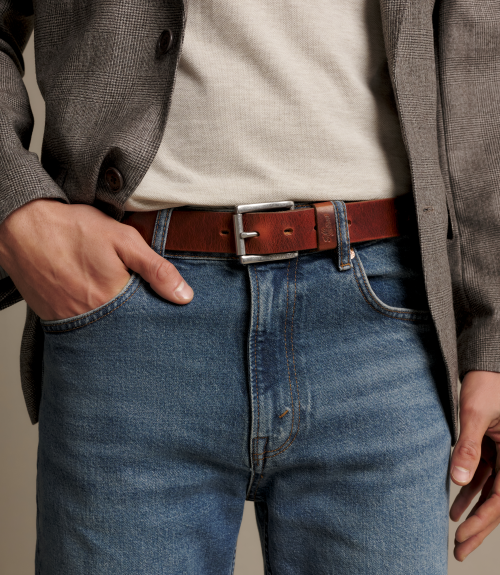 A close-up of a man wearing a brown leather belt with a silver buckle, styled with blue jeans, a beige t-shirt, and a gray blazer.