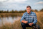 Man in a blue plaid shirt sitting by a serene lakeside, surrounded by tall grass and nature.