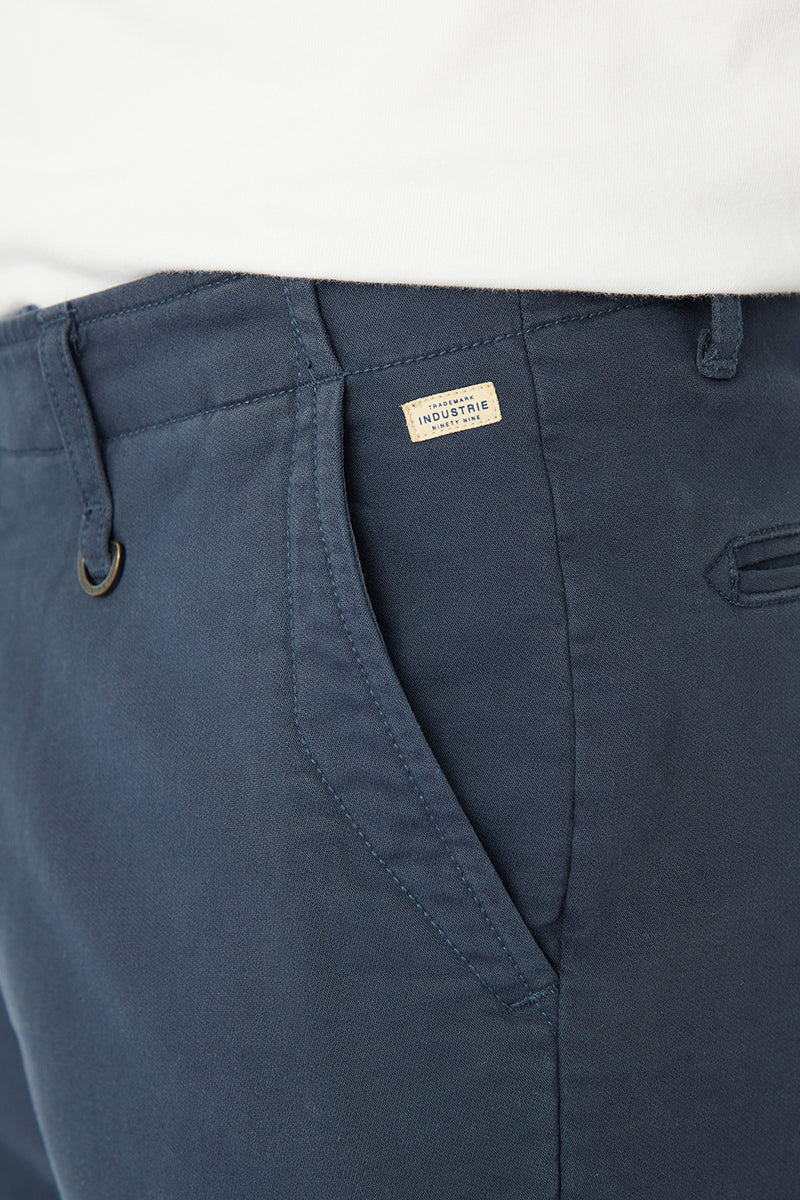 Close-up of navy blue chino pants showing the waistband, belt loops, front pocket, and a small beige brand label that reads 'The Normal Brand'.