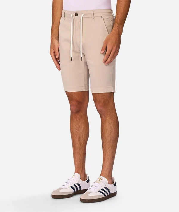 A person wearing light beige drawstring shorts, a pale pink top, and white sneakers with dark stripes, shown from the waist down against a light gray studio background.