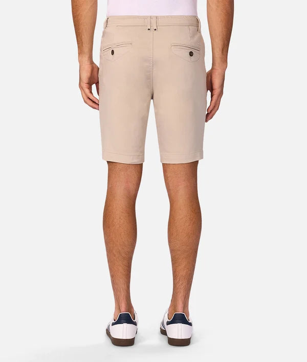 A man viewed from the back wearing light beige chino shorts, a white shirt, and white loafers with navy accents, set against a plain light gray studio background.