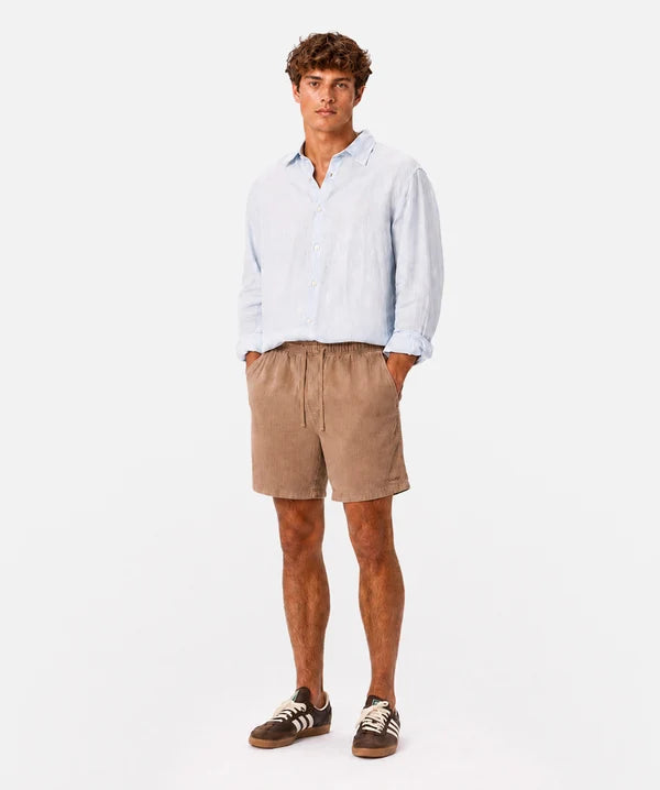 A man modeling a light blue long-sleeve linen shirt and tan corduroy shorts against a plain white studio background.