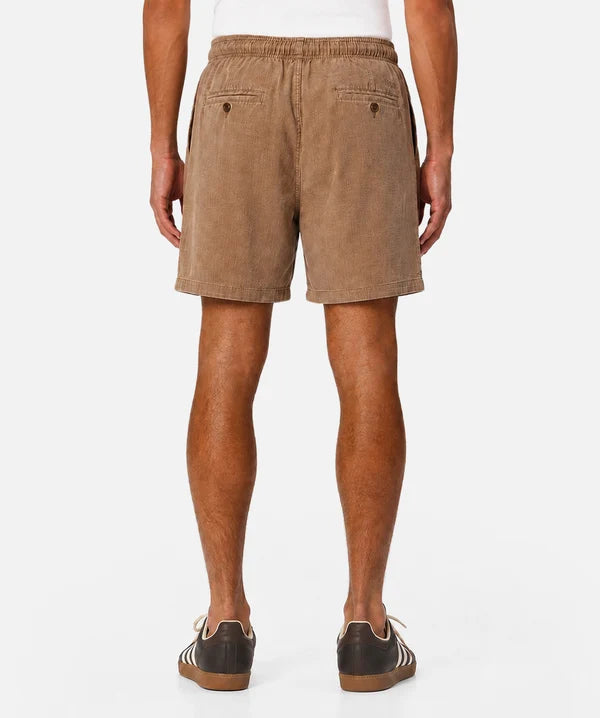 A person shown from the back wearing tan corduroy shorts, a white top, and dark brown sneakers with white stripes, against a plain light gray studio background.