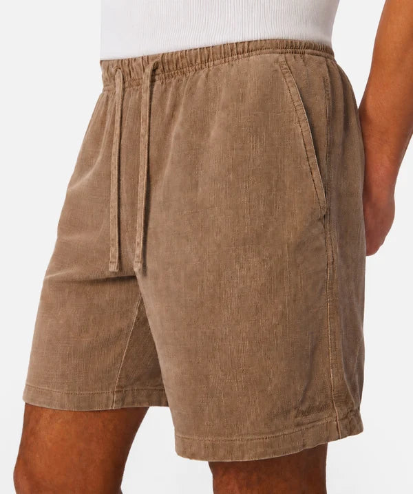 A close-up of a person wearing brown corduroy shorts with an elastic waistband and drawstring, paired with a white ribbed top against a plain light background.