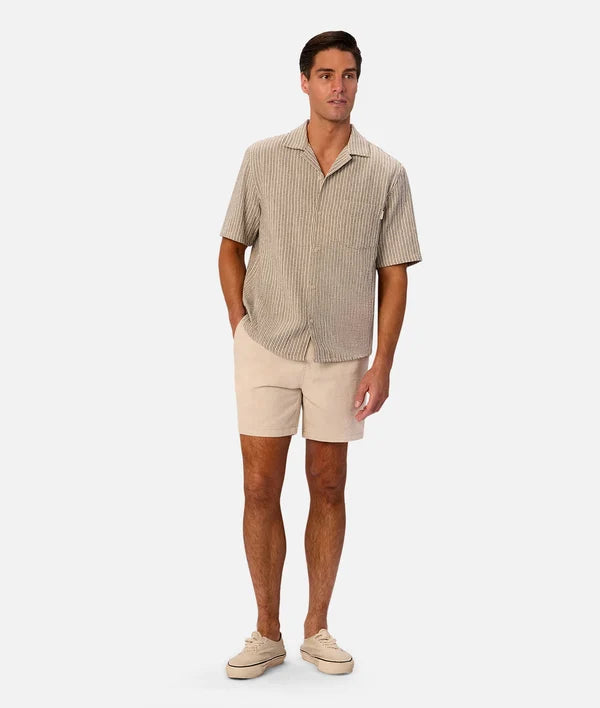 A man modeling a short-sleeve button-up shirt with thin vertical stripes in beige and brown, paired with beige shorts and beige sneakers, against a light gray studio background.