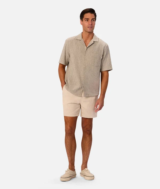 A man modeling a short-sleeve button-up shirt with thin vertical stripes in beige and brown, paired with beige shorts and beige sneakers, against a light gray studio background.