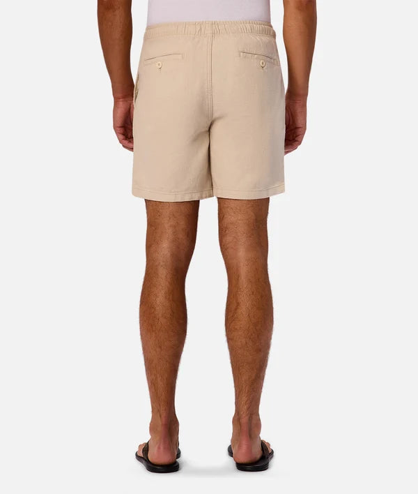 A man viewed from the back wearing light beige chino shorts, black flip-flops, and a white t-shirt against a light gray studio background.