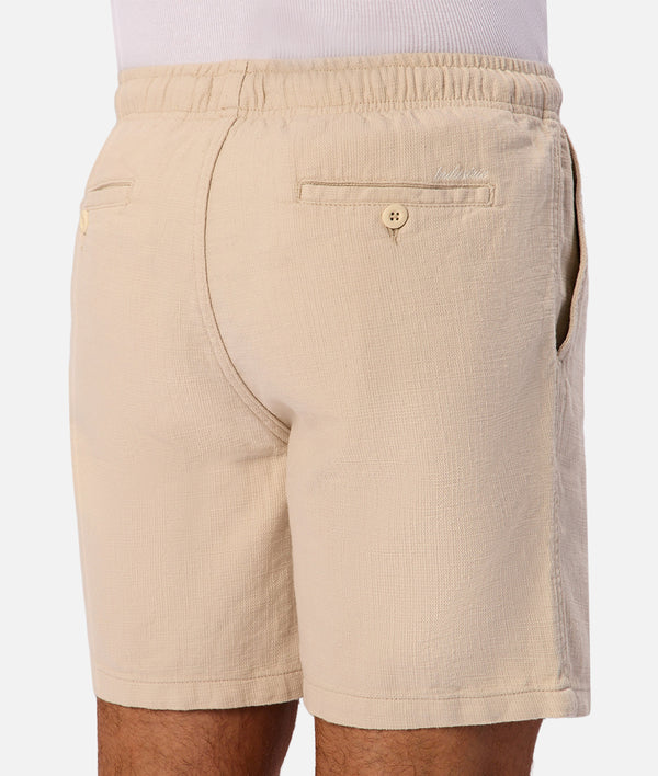 Beige textured cotton shorts with an elastic waistband, shown from the back on a model.