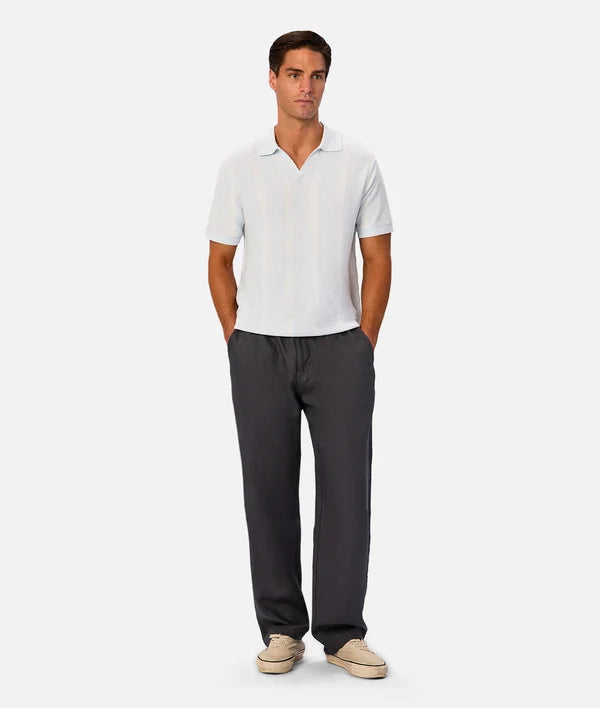 A man modeling a white short-sleeve polo shirt and dark gray drawstring trousers with beige slip-on shoes, standing against a light gray studio background.