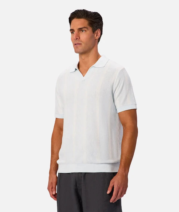 A man wearing a light blue short-sleeve knit polo shirt and dark gray trousers, standing against a solid light gray background.
