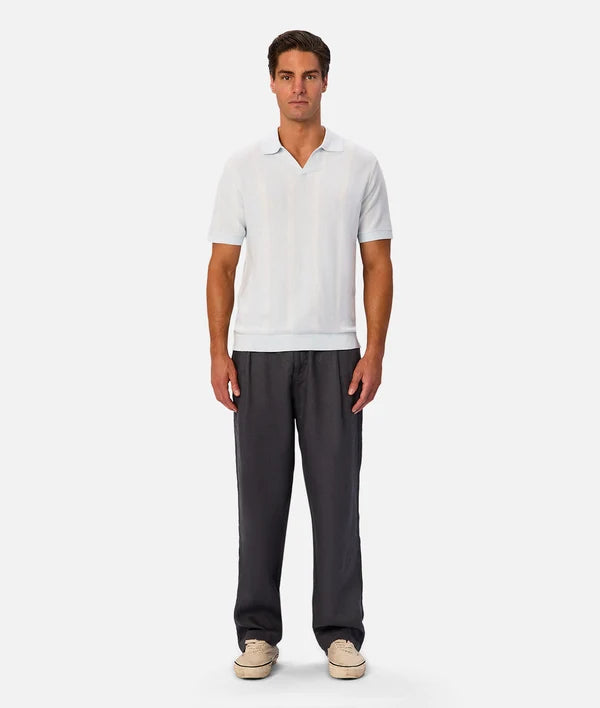 A man modeling a white short-sleeve polo shirt paired with dark gray trousers and beige sneakers, standing against a light gray studio background.