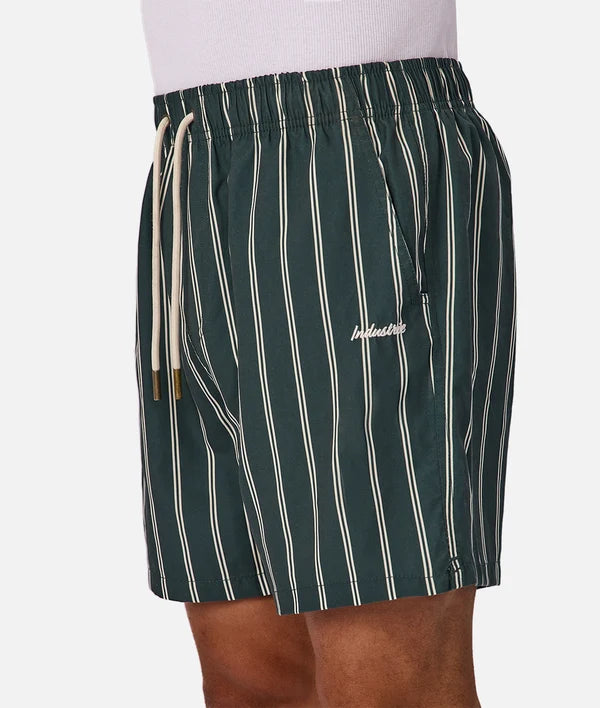 A close-up of a person wearing dark green drawstring shorts with thin vertical cream stripes and a small white script logo on the left leg, against a light gray studio background.