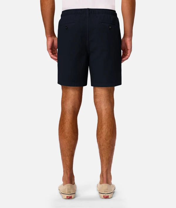 A man viewed from the back wearing dark navy blue shorts, a white t-shirt, and beige slip-on shoes against a light gray studio background.