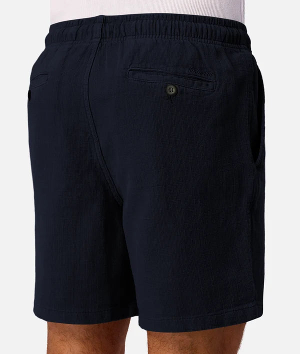 Close-up rear view of a person wearing dark navy shorts with an elastic waistband, side pockets, and a buttoned back pocket against a light gray studio background.