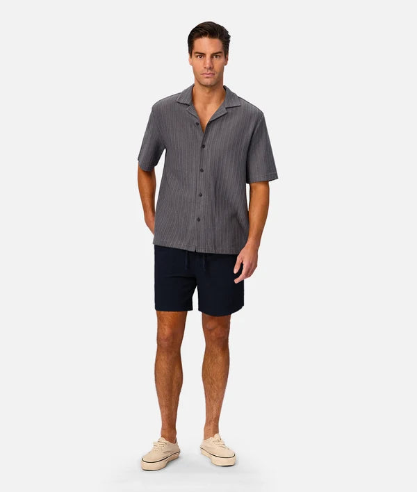 A man modeling a short-sleeve button-up shirt with a fine vertical stripe pattern in gray and black, paired with dark navy shorts and beige sneakers, against a light gray studio background.