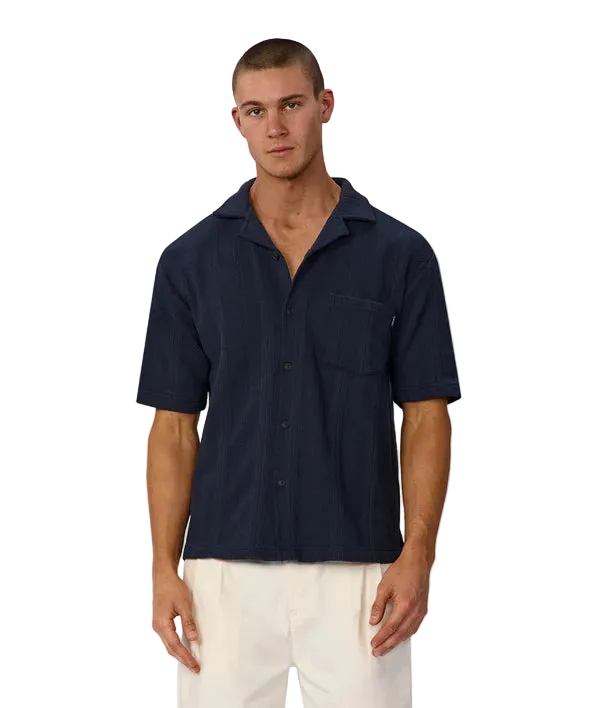 A man wearing a dark navy blue short-sleeve button-up shirt with a chest pocket, paired with off-white pants, standing against a light gray background.