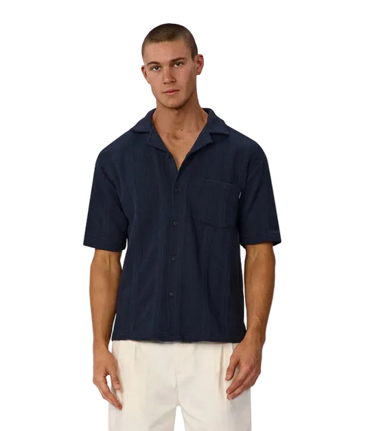 A man wearing a dark navy blue short-sleeve button-up shirt with a chest pocket, paired with off-white pants, standing against a light gray background.