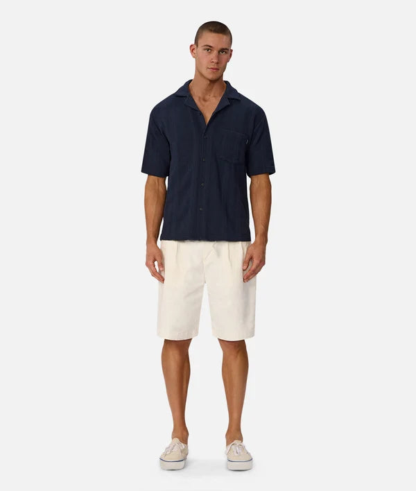 A male model stands against a light gray studio background wearing a dark navy short-sleeve button-up shirt, off-white shorts, and white sneakers.
