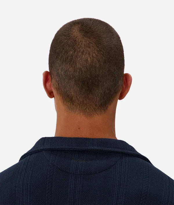 Back view of a man with a buzz cut wearing a dark navy textured knit polo shirt against a light gray background.