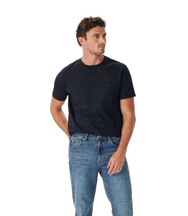 A man wearing a dark navy blue crewneck t-shirt with a small red embroidered logo on the left chest, paired with blue jeans, photographed against a solid white background.