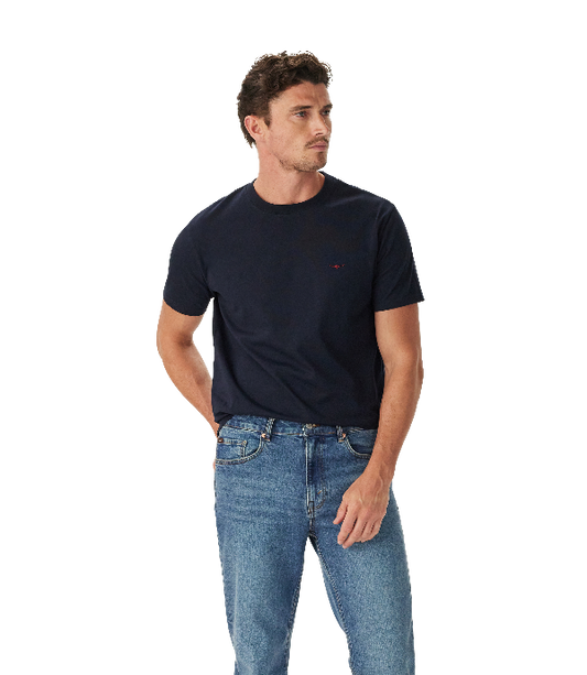 A man wearing a dark navy blue crewneck t-shirt with a small red embroidered logo on the left chest, paired with blue jeans, photographed against a solid white background.