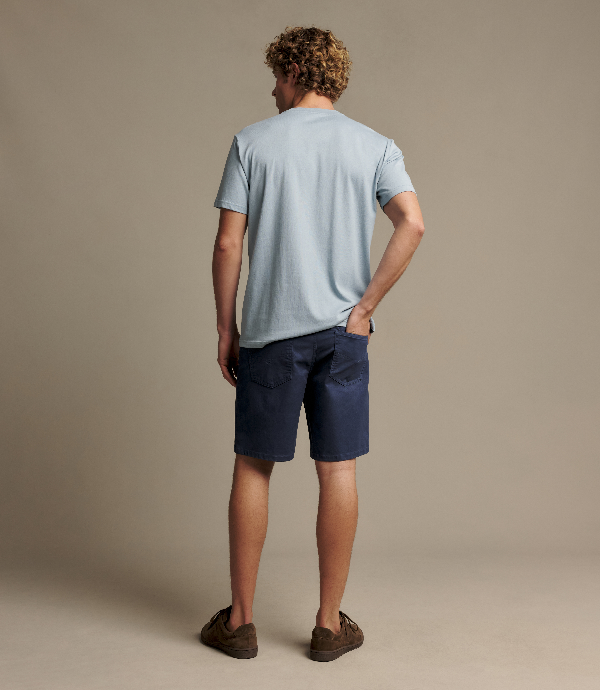 A man standing with his back to the camera wearing a light blue t-shirt, navy blue shorts, and brown loafers, photographed in a studio setting against a solid beige background.