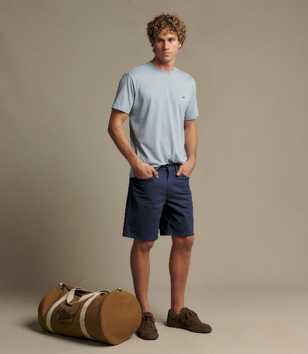 A man stands in a studio wearing a light blue crew-neck t-shirt, navy shorts, and brown shoes, with a brown duffel bag on the floor beside him against a warm beige background.