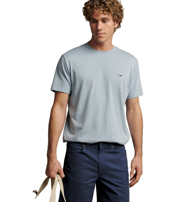 A man wearing a light blue crewneck t-shirt with a small embroidered bird logo on the chest, paired with navy blue pants, holding a beige tote bag against a solid beige background.