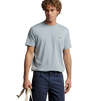 A man wearing a light blue crewneck t-shirt with a small embroidered bird logo on the chest, paired with navy blue pants, holding a beige tote bag against a solid beige background.