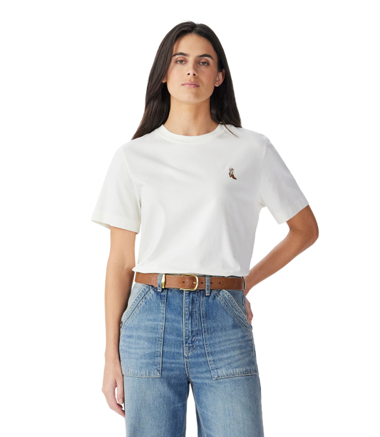A woman modeling a white short-sleeve crewneck t-shirt with a small embroidered polo player logo on the left chest, paired with a brown belt and blue jeans against a plain white background.
