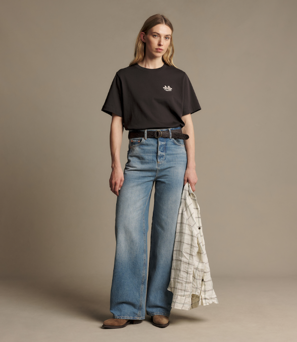 A model stands against a plain beige studio background wearing a black short-sleeve T-shirt with a small white chest logo, wide-leg blue jeans, a black belt, and holding a light plaid shirt under one arm.